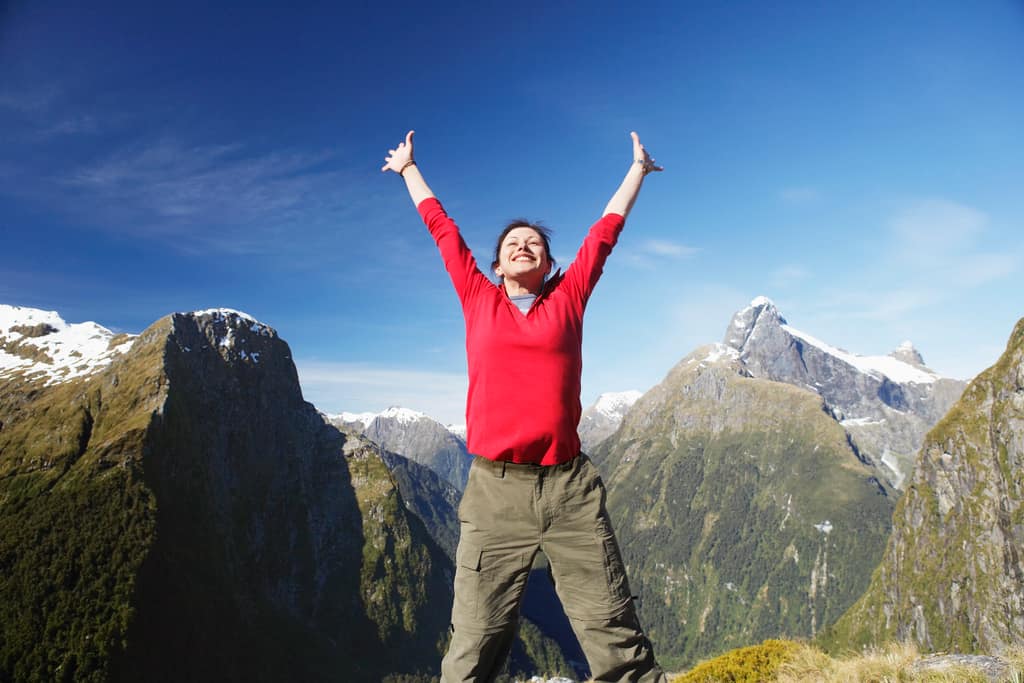 Man in a red top celebrating with arms raised in the mountains